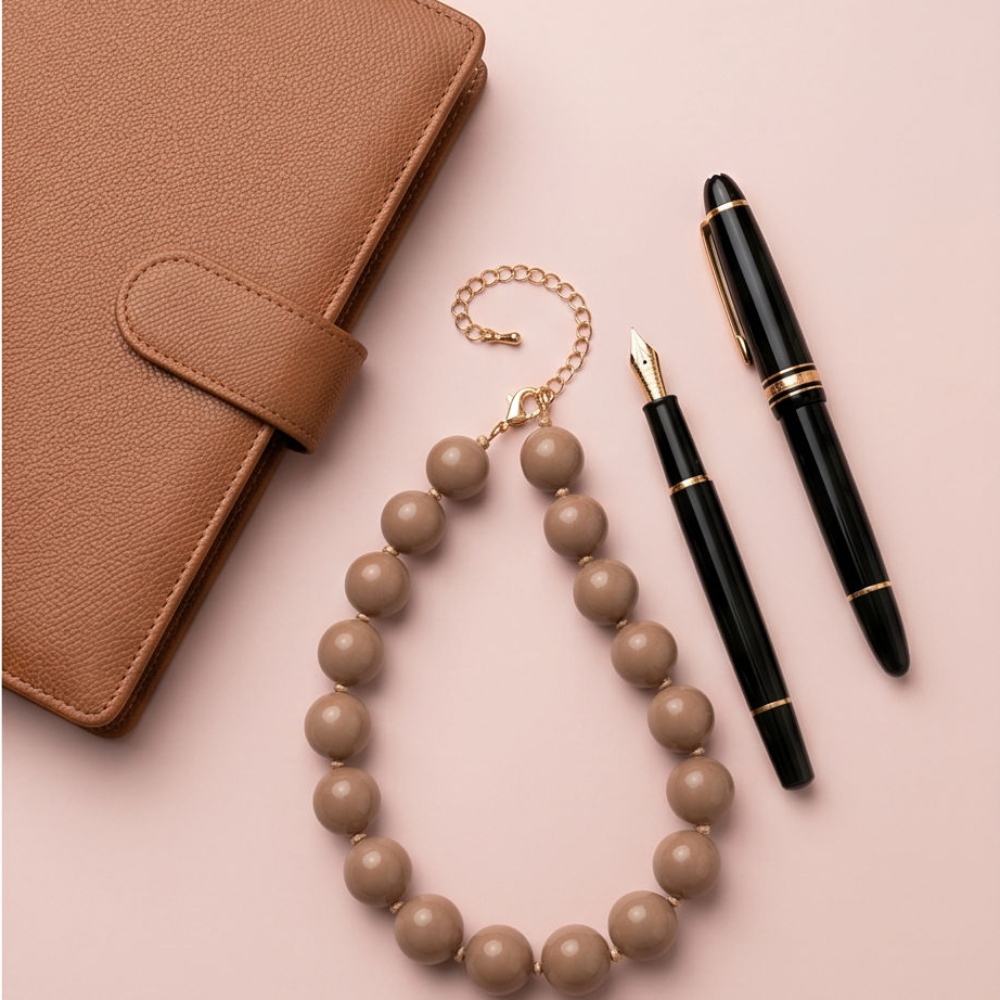 A styled flat-lay featuring a tan leather planner cover, a black fountain pen with gold trim, and a matching tan beaded necklace with a gold chain link, arranged on a solid pale pink background.